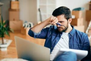 Distraught man reading an e-mail on laptop while moving into a new home.