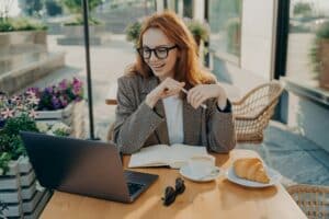 Young smiling woman sitting at cafe table outdoors and watching online lesson or webinar on laptop